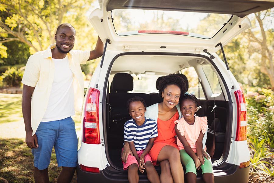 About Our Agency - Family Sits in the Back of Their White Car, Smiling, All Dressed for Warm Weather and Parked Beneath Trees